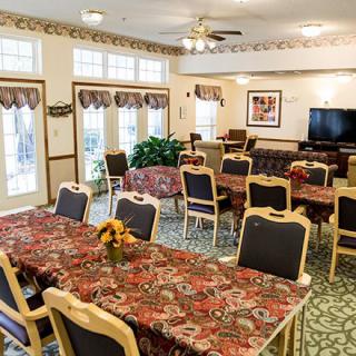 A well-lit dining area with patterned tablecloths, multiple chairs, a television, and large windows.
