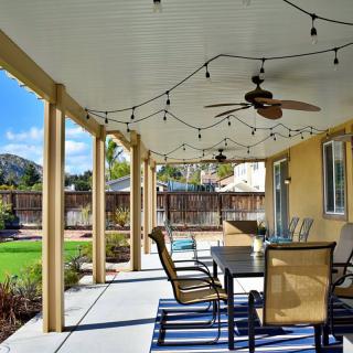 A charming covered patio with outdoor seating, string lights, and a ceiling fan overlooking a sunny backyard.