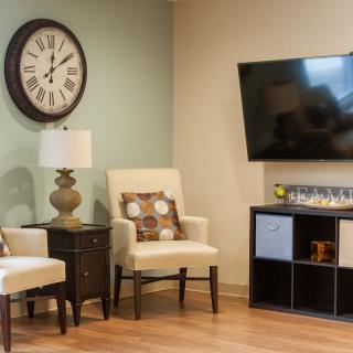 A cozy living room with a large clock, comfortable armchairs, and a mounted television.