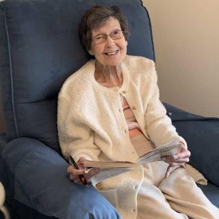 An elderly woman smiling while seated in a chair, holding a book.