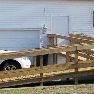A wooden accessibility ramp leads to the entrance of a house next to a garage with a parked car.