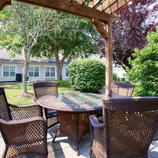 A cozy outdoor patio area featuring a round table with wicker chairs under a pergola, surrounded by lush greenery.