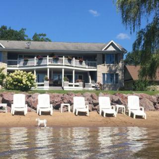 A lakefront house with a row of white lawn chairs on the sandy beach.