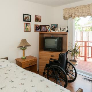 A cozy bedroom featuring a bed, a wheelchair beside a dresser with an old television, and family photos on the wall.