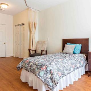 A cozy bedroom featuring a single bed with patterned bedding and decorative pillows, complemented by wooden flooring, an armchair, and a nightstand with a lamp.