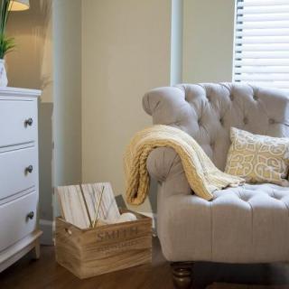A cozy living room corner with a tufted armchair, soft blanket, and decorative pillow beside a wicker basket and dresser.