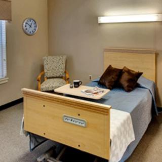 A clean and well-organized hospital room featuring a bed with pillows, a small table with a cup and magazine, and a comfortable chair.