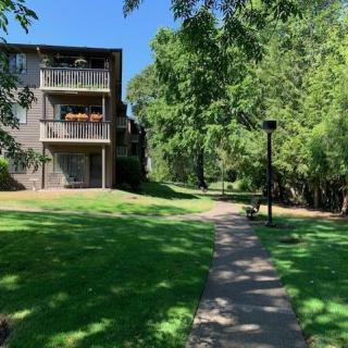 A serene outdoor scene with a paved pathway leading past an apartment building, surrounded by lush green trees and grass.