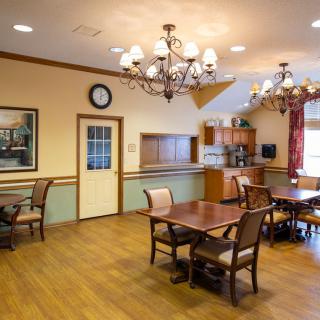 A cozy dining room with wooden flooring and elegant chandeliers, featuring neatly arranged chairs and tables.