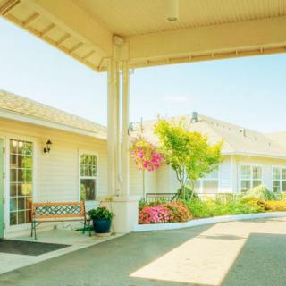A sunny outdoor area with a building featuring a covered entrance, landscaped flowers, and a bench.