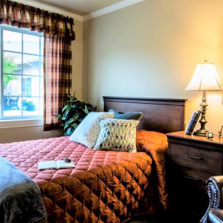A cozy bedroom with a neatly made bed, a lamp on the nightstand, and sunlight coming through the window.