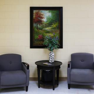 A cozy waiting area featuring two gray chairs, a small table with a decorative vase, and a nature-themed painting on the wall.