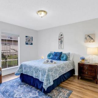 A cozy bedroom with a double bed, featuring blue and white bedding, surrounded by wall art and a nightstand with a lamp.