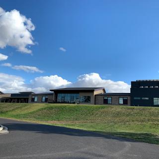 A modern building set against a bright blue sky with scattered clouds, surrounded by grass and a paved road.