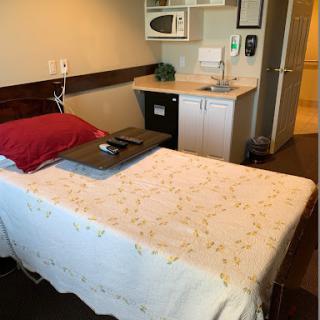 A patient room featuring a bed with a red pillow, a small kitchenette with a microwave, and a table.