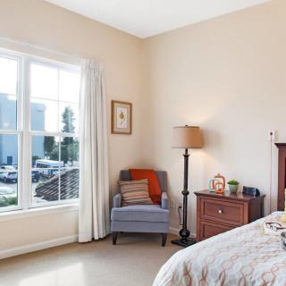 A cozy bedroom featuring a comfortable chair by the window and a bedside lamp.