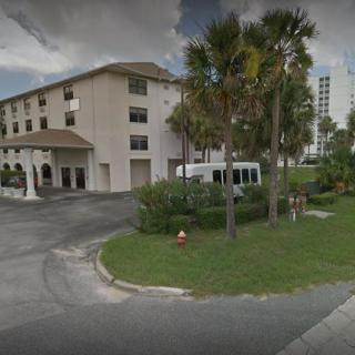 A three-story building with palm trees and a vehicle parked nearby under a cloudy sky.
