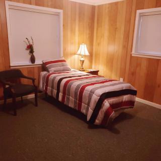 A cozy bedroom with wood-paneled walls, featuring a single bed with striped bedding, a lamp on a side table, and a chair in the corner.