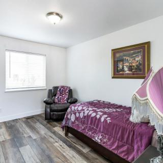 A cozy bedroom with a single bed draped in purple bedding, an armchair beside it, a decorative lamp, and a painting on the wall.