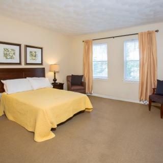 A cozy bedroom with a yellow bedspread, framed art on the wall, and two windows with light curtains.