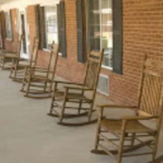 A row of wooden rocking chairs lined up on a porch in front of a brick building.