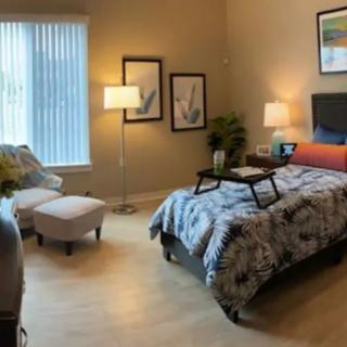 A cozy, modern bedroom interior featuring a neatly made bed with printed bedspread, accent chairs, soft lighting, and framed artwork on the walls.