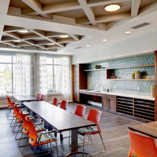 A modern conference room with a long table and vibrant orange chairs, featuring large windows and a stylish ceiling design.
