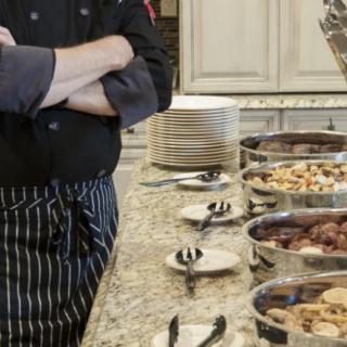 A chef standing beside a buffet setup with various dishes and plates in a kitchen setting.