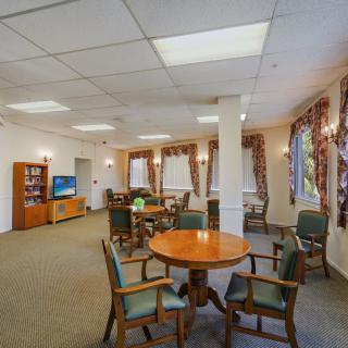 A cozy living area featuring round tables, chairs, a television, and large windows with drapes.