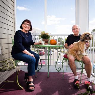 A couple sits on their porch with a dog, enjoying a casual daytime moment.