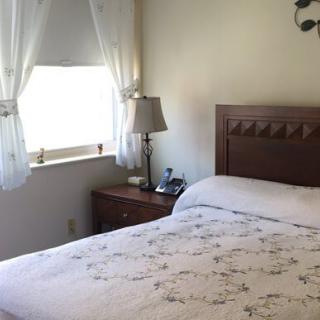 A cozy bedroom with a neatly made bed, a wooden nightstand with a lamp, and light curtains covering the window.