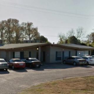 A small building with a parking lot filled with cars, surrounded by trees on a sunny day.