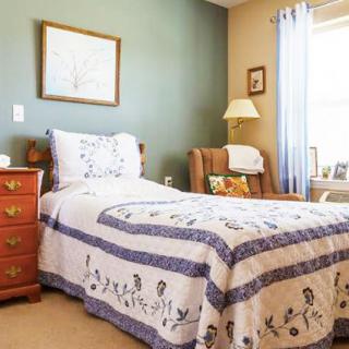 A cozy bedroom featuring a single bed with a floral quilt, a wooden drawer, and a comfortable armchair by the window.