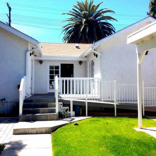 A white house with a ramp leading to the entrance and a well-maintained lawn, framed by a tall palm tree.
