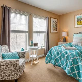 A cozy bedroom with a teal and white patterned bedspread, featuring an armchair, natural light from two large windows, and decorative artwork on the walls.