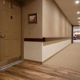 A well-lit hotel hallway with a wooden floor and carpet, featuring a closed door on the left.