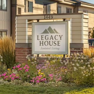 A sign for Legacy House Assisted Living surrounded by a garden with flowers in front of the building.