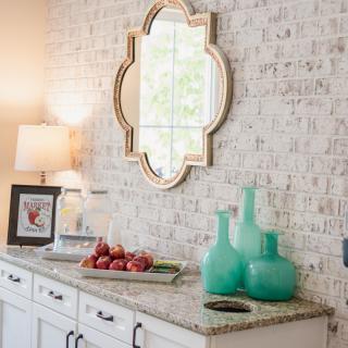 A stylish interior features a sideboard with decorative vases, a lamp, and a unique mirror on a brick wall.