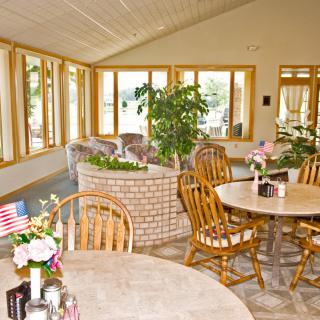 A cozy dining room with wooden tables and chairs, surrounded by large windows and indoor plants.