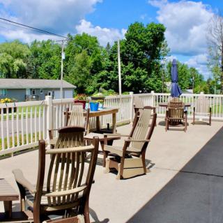 A sunny patio with wooden chairs and tables, surrounded by a white fence and trees in the background.