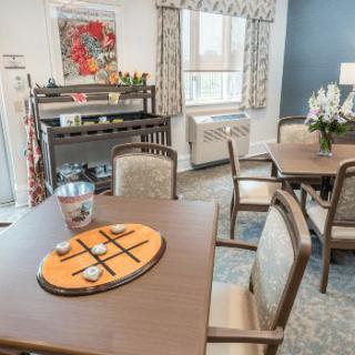 A cozy dining room with neatly arranged tables and chairs, featuring a decorative tic-tac-toe game and a vase of flowers.