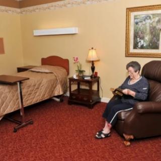 An elderly woman reads in a comfortable chair within a nursing home bedroom setting.