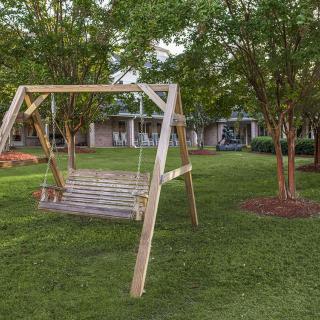 A wooden swing bench in a lush garden setting surrounded by trees and green grass.