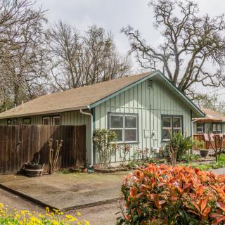 A quaint cottage surrounded by trees and shrubs, featuring a small garden and patio area.