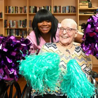 An elderly person and a woman posing with colorful pom poms in a library.