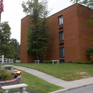 A brick building surrounded by trees and grass, with a pathway and benches in front.