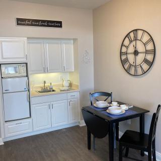 A compact kitchen space with white cabinets, a small dining table set for two, and a large wall clock.