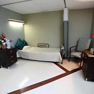 A hospital room featuring a bed, nightstand, and chair, with soft lighting.