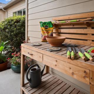 A wooden potting bench is set up with gardening tools and a watering can, surrounded by potted plants.