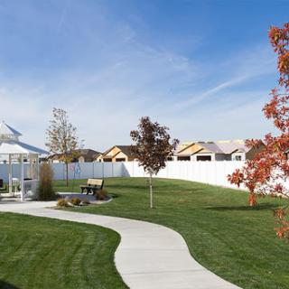 A serene garden pathway winding through a well-maintained lawn, with vibrant trees and houses in the background under a clear blue sky.